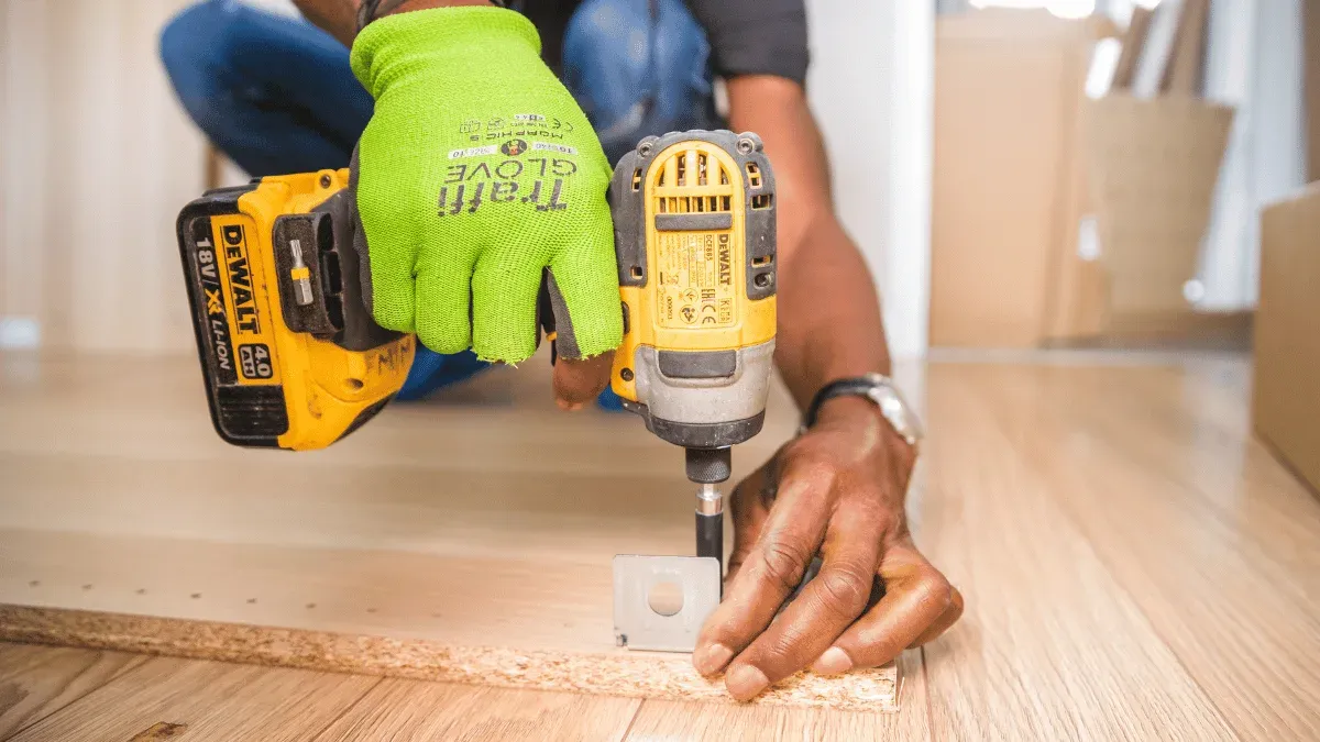 Mains gantées d’un artisan qui visse une ferrure sur un panneau de bois avec une visseuse sans fil